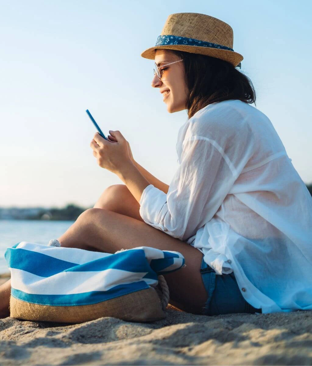 Woman in straw hat reading phone at beach. Download your free ebook. Real Estate Investing for beginners