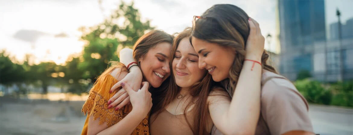 Three sisters embracing with a blurred high-rise backdrop on a sunny day, reflecting the resolution of probate property disputes. Reach out to Kid Breukelen Realty Group for professional help in resolving property issues.