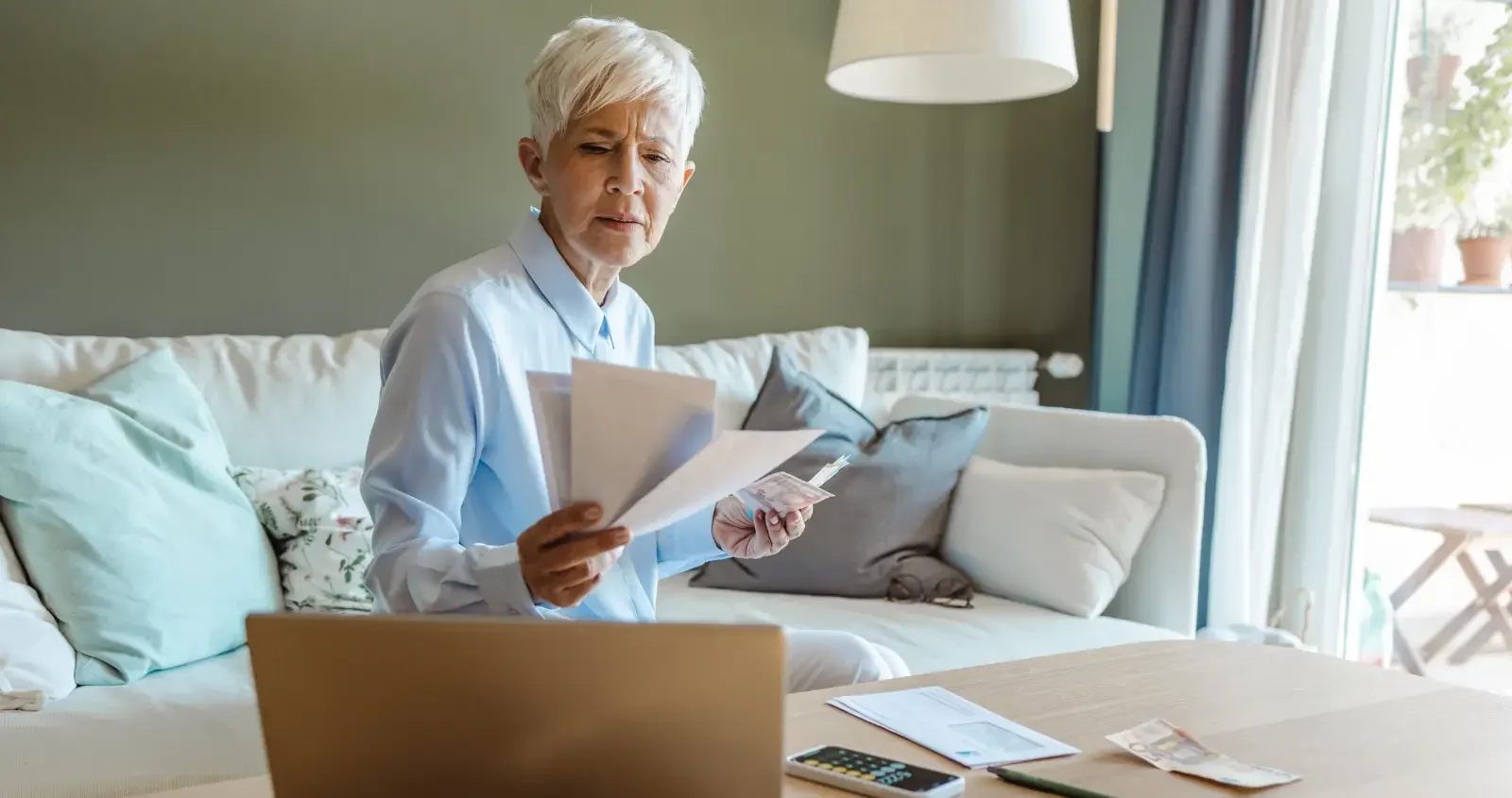 A woman sitting on a couch near a window reviewing bills with a laptop open, representing mortgage or foreclosure-related property stress in Broward.